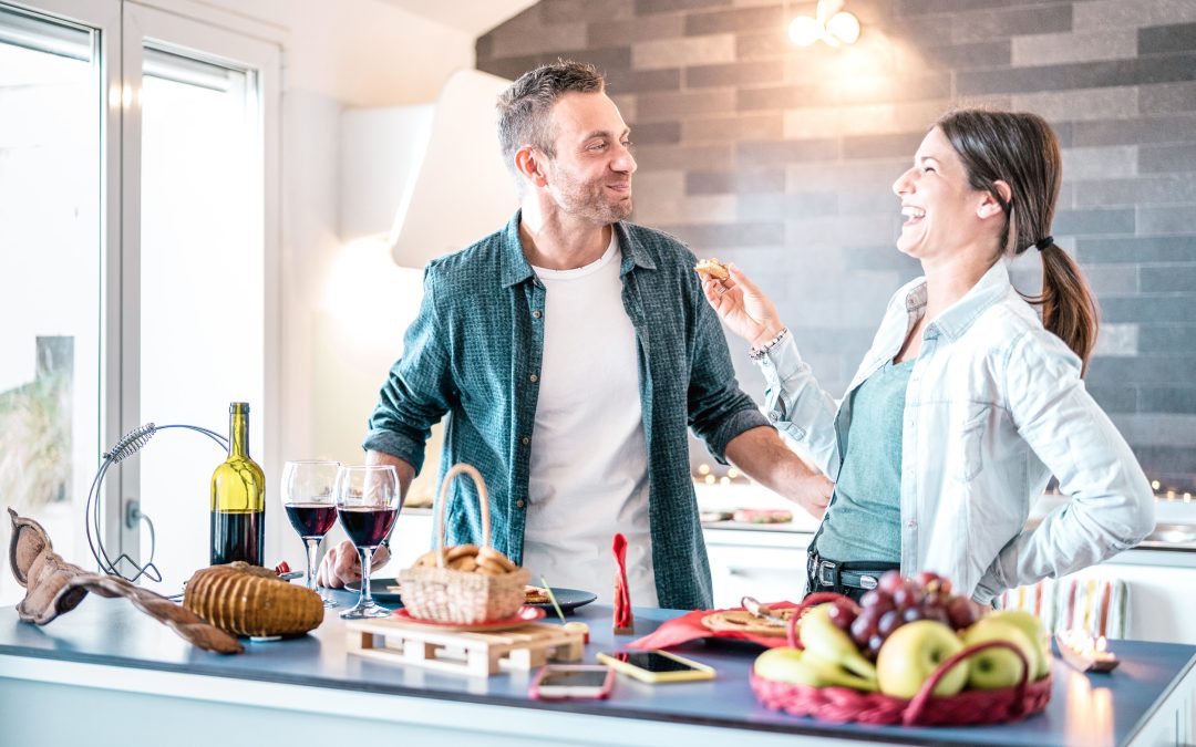 Couple enjoying food and wine in a modern kitchen, featuring a variety of culinary supplies and fresh fruits, highlighting the joy of home cooking.