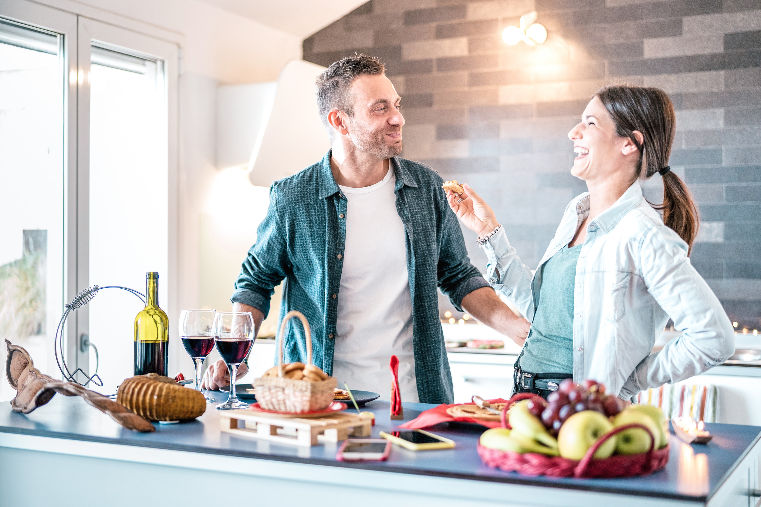 Young couple in love having fun at house kitchen - Happy milleni Couple enjoying food and wine in a modern kitchen, featuring a variety of snacks, wine glasses, and fresh fruits, highlighting a warm and inviting culinary experience.